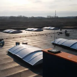 Płaski dach przemysłowy z świetlikami kopułowymi i elementami wentylacji, widok z góry w słoneczny dzień. Na horyzoncie drzewa i błękitne niebo.