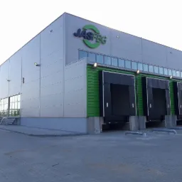 Nowoczesna hala magazynowa JASFBG z szarymi panelami i zielonymi akcentami, widoczne doki załadunkowe z czarnymi bramami, betonowy plac przed budynkiem.