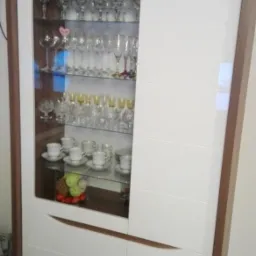 Nowoczesna witryna z białymi frontami i drewnianą ramą, wypełniona szkłem, eksponująca kolekcję kieliszków, filiżanek i dekoracyjnych kul.