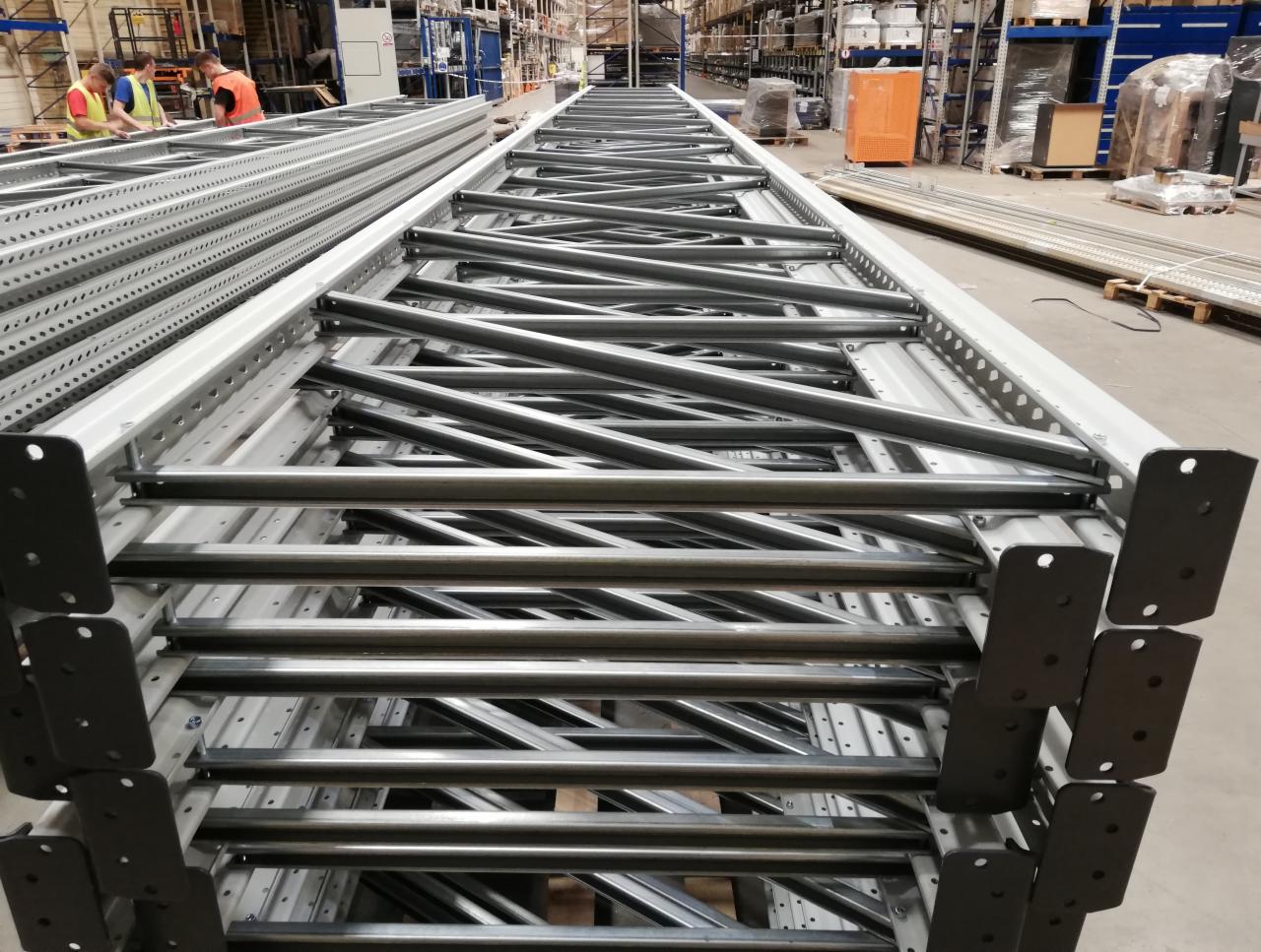 Stos metalowych, perforowanych słupów regałowych z poprzecznymi wzmocnieniami, ułożonych jeden na drugim w magazynie, widoczni pracownicy w tle.