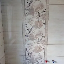 Beżowe płytki ścienne z kwiatowym wzorem w pionowym pasie, widoczne przyłącza wodne z czerwonym i niebieskim oznaczeniem na tle wanny.