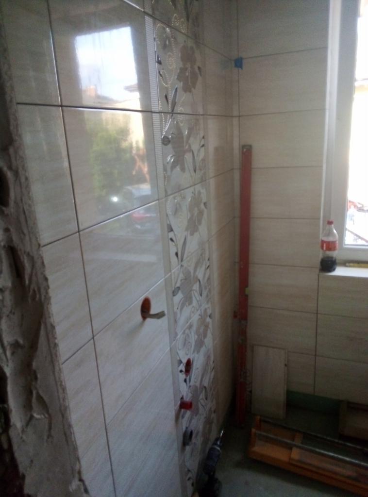 Układanie beżowych płytek z motywem kwiatowym na ścianie łazienki, widoczne otwory na instalacje, poziomica oparta o ścianę, w tle okno i butelka z napojem.