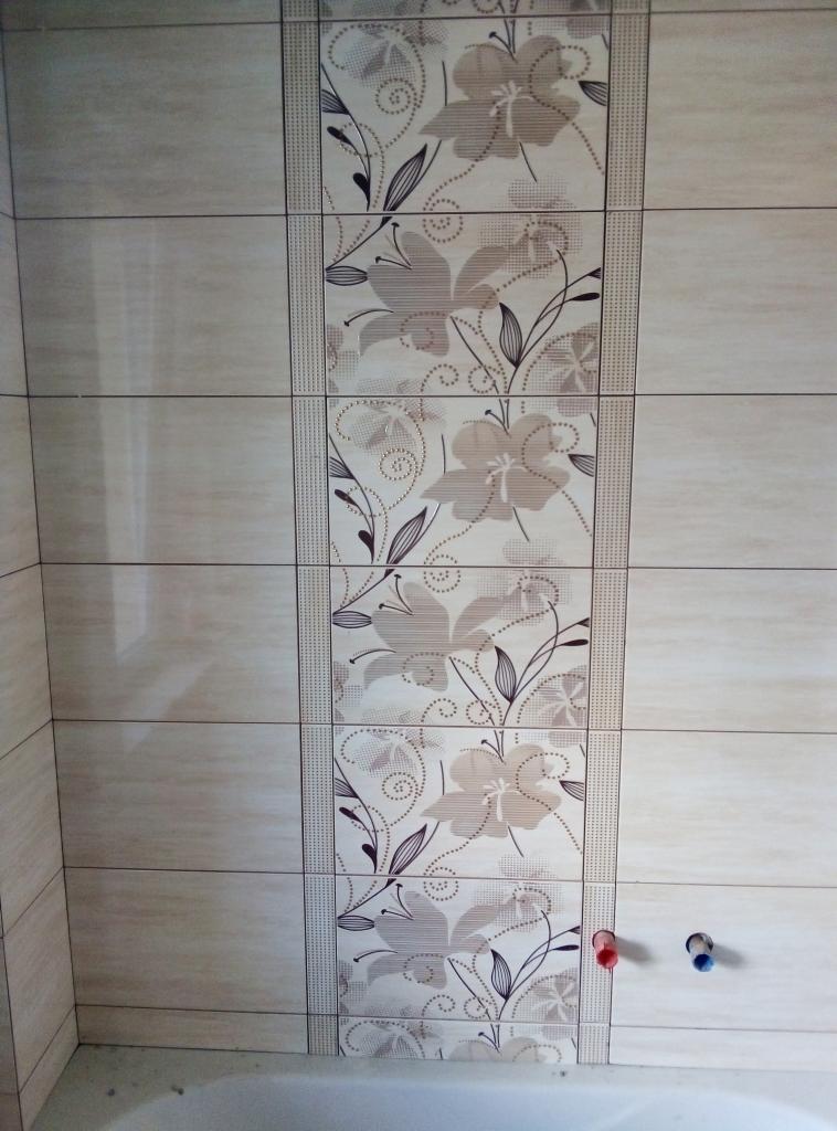 Beżowe płytki ścienne z kwiatowym wzorem w pionowym pasie, widoczne przyłącza wodne z czerwonym i niebieskim oznaczeniem na tle wanny.