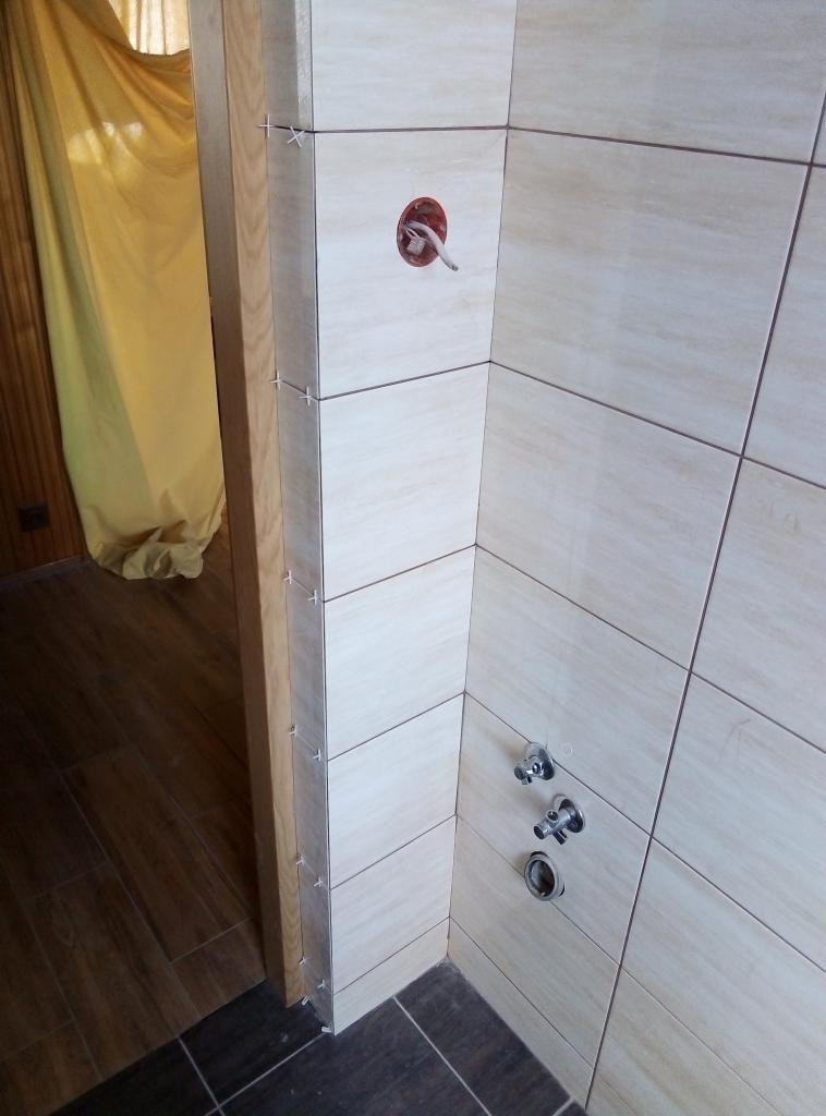 Świeżo ułożone, beżowe płytki ceramiczne na ścianie łazienki, z widocznymi krzyżykami dystansowymi i przygotowanymi otworami na armaturę, kontrastujące z ciemnymi płytkami podłogowymi i drewnianą...