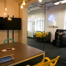 Nowoczesne biuro z salą konferencyjną, laptopem na stole i widokiem na strefę relaksu z sofą i neonem. Minimalistyczny design, jasne kolory, przestronność.