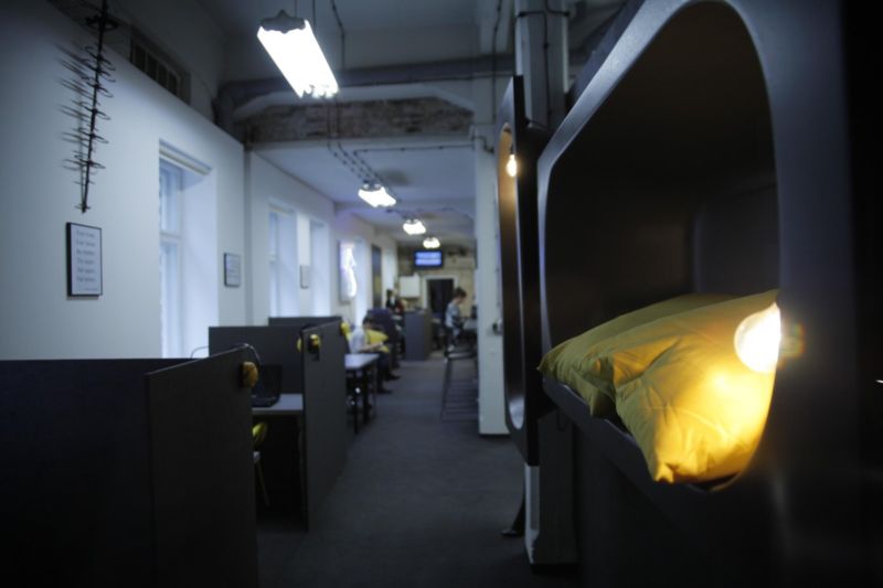 Nowoczesne wirtualne biuro z kapsułami do spania, oświetlone lampami. W tle widoczne stanowiska pracy i osoby. Unikalny design wnętrza.