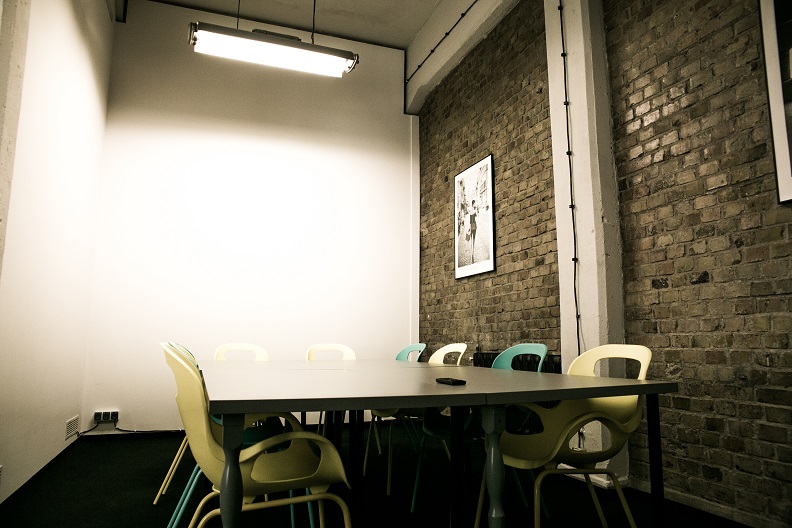 Sala konferencyjna z długim stołem i designerskimi krzesłami na tle ceglanej ściany z plakatem. Minimalistyczne oświetlenie, surowy, industrialny styl wnętrza biurowego.