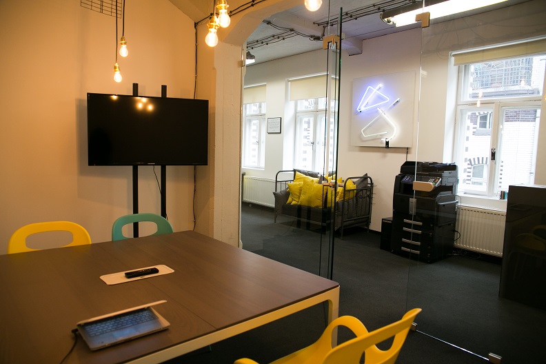 Nowoczesne biuro z salą konferencyjną, laptopem na stole i widokiem na strefę relaksu z sofą i neonem. Minimalistyczny design, jasne kolory, przestronność.