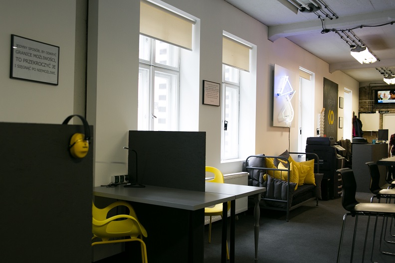 Nowoczesne wirtualne biuro: żółte krzesła, szare biurka, kanapa z poduszkami. Na ścianie plakat z motywacyjnym hasłem i neonowa grafika. Okna z roletami w tle.