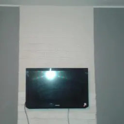 Ściana pomalowana na dwa kolory: szary i biały imitujący cegłę. Na białej części wisi telewizor. Widoczny fragment sufitu i podłogi.