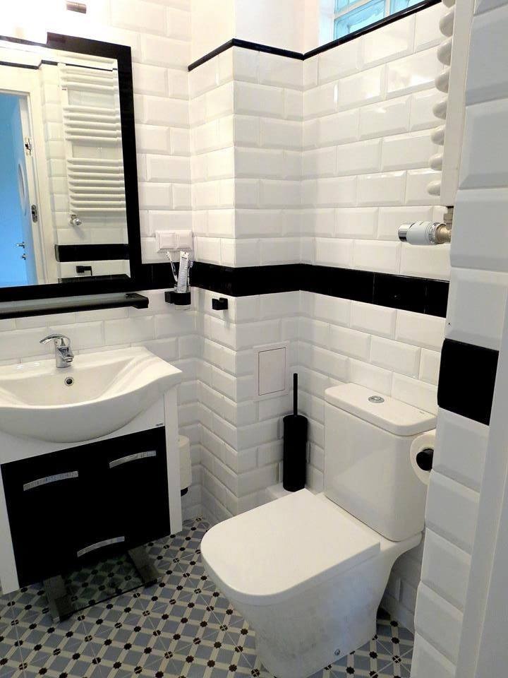 Biała łazienka z czarnymi akcentami: umywalka z szafką, toaleta i białe kafelki typu metro z czarnym pasem. Na podłodze kafelki w geometryczny wzór.
