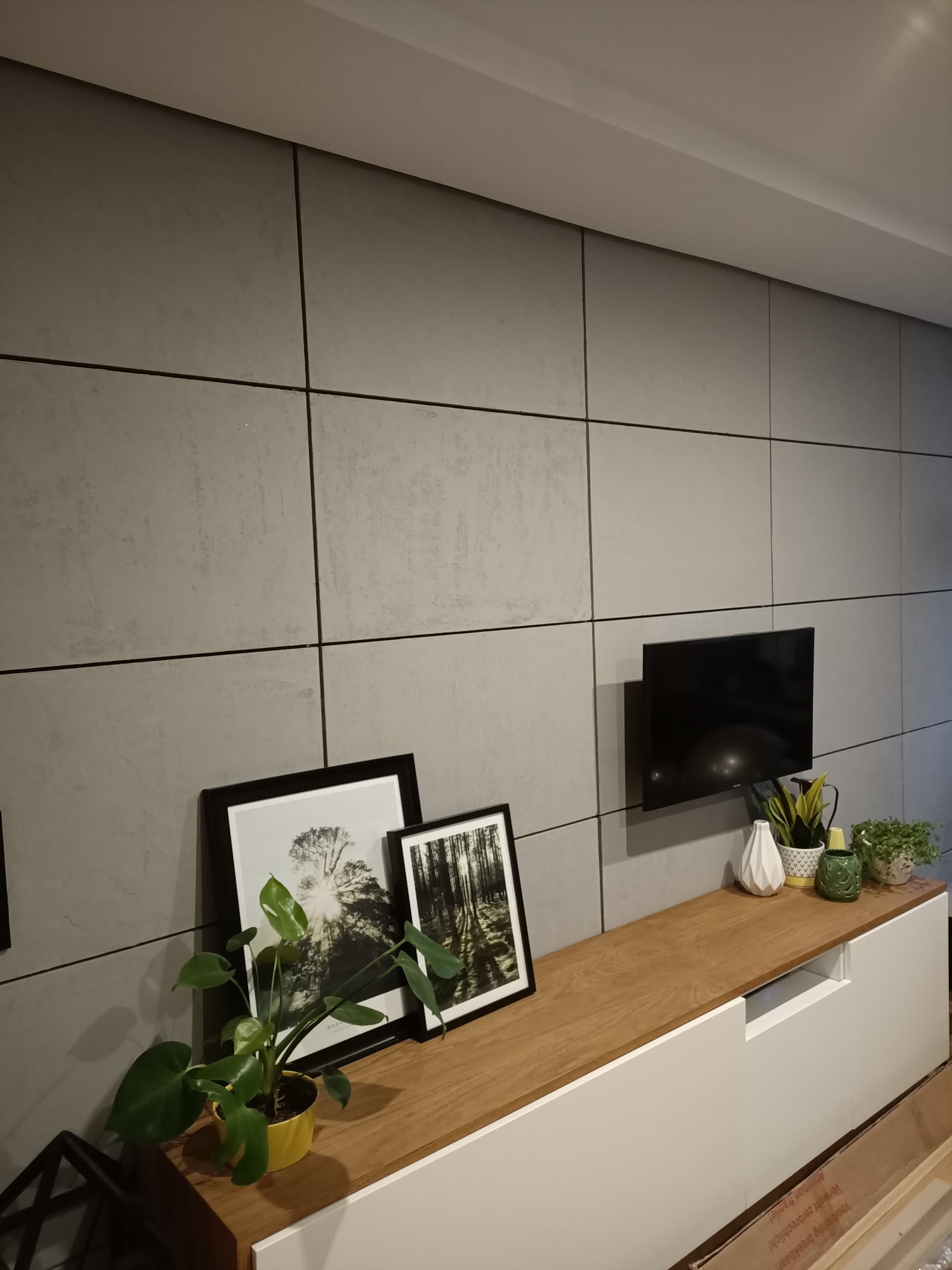 Szara ściana wykończona panelami z mikrocementu z czarnymi fugami, podwieszony telewizor, drewniany blat szafki RTV z dekoracjami: ramki ze zdjęciami, biały wazon i doniczki z roślinami.