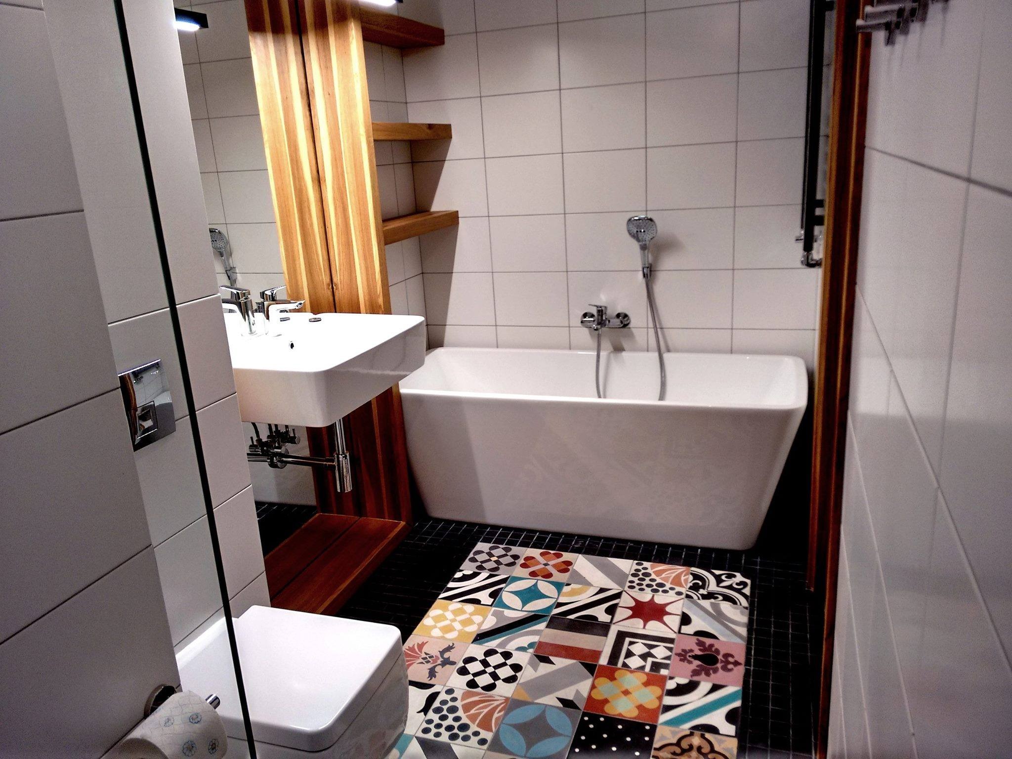 Nowoczesna łazienka z białą wanną, umywalką i mozaikową podłogą, akcentowana drewnianymi elementami i białymi płytkami.