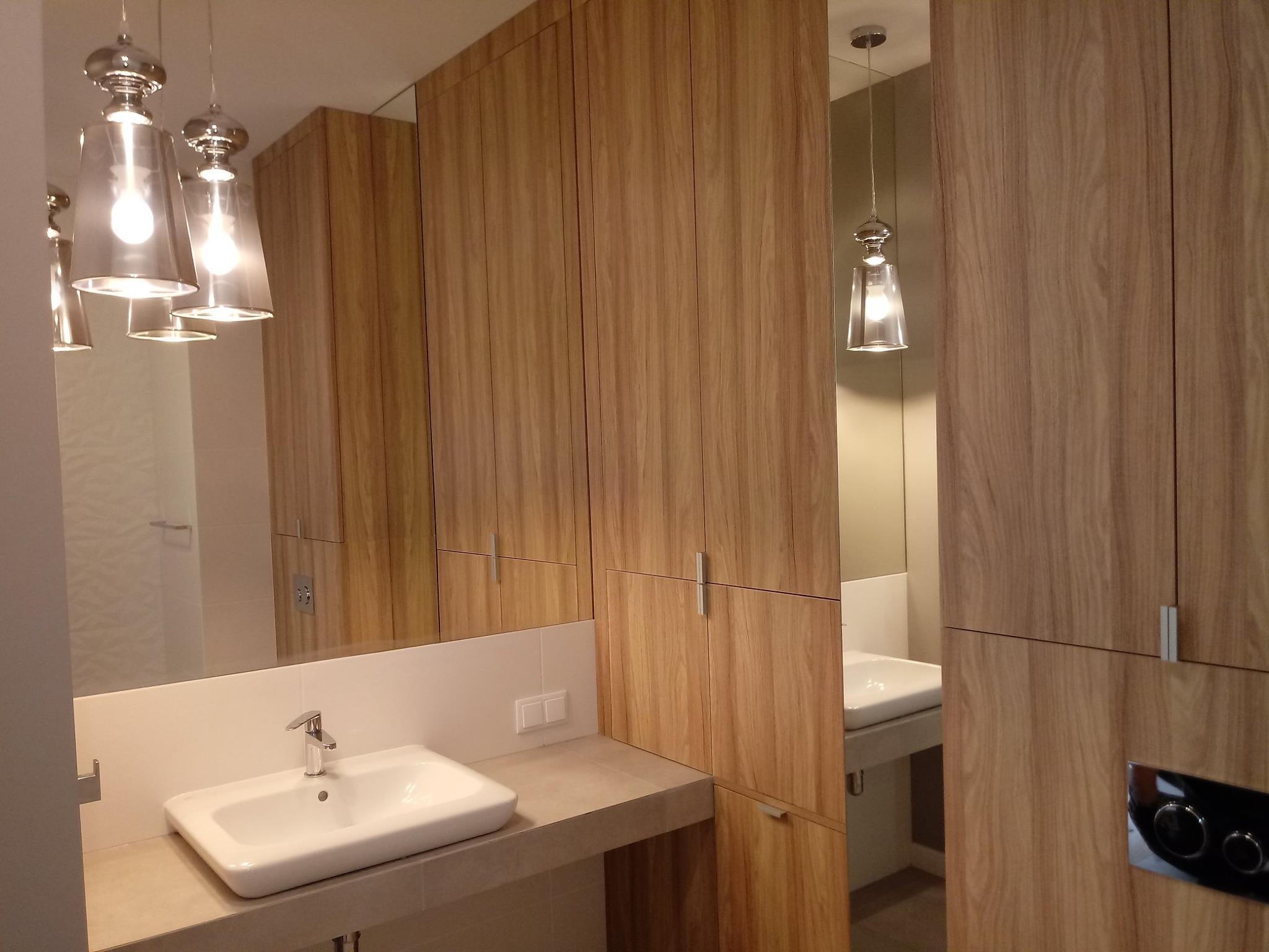 Nowoczesna łazienka z umywalką nablatową, armaturą, lustrem i zabudową z drewnianych szafek z podłużnymi uchwytami, oświetlona wiszącymi lampami z przezroczystymi kloszami.