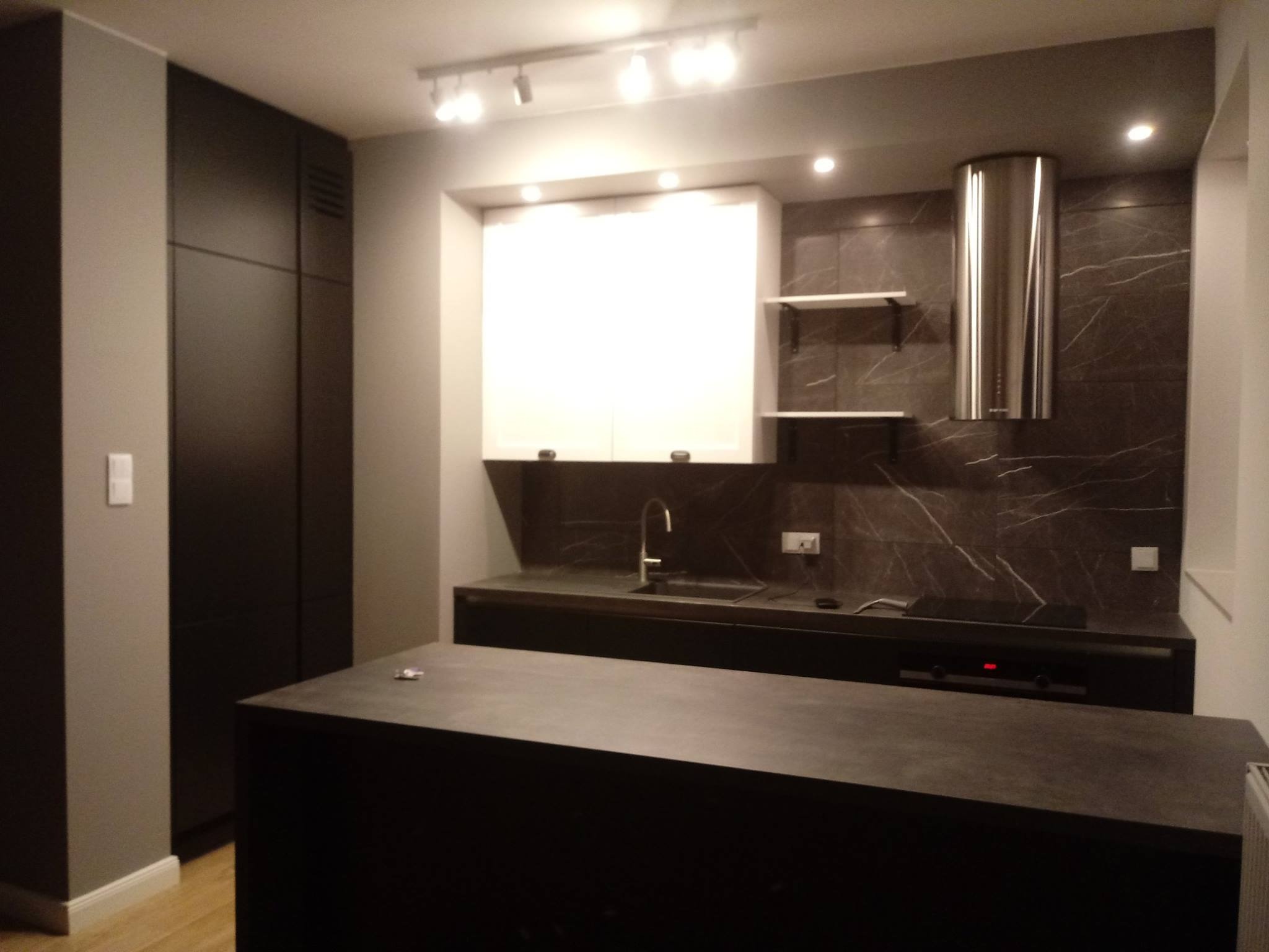 Nowoczesna kuchnia w odcieniach szarości i czerni z wyspą kuchenną, białymi szafkami wiszącymi i marmurową ścianą nad blatem.