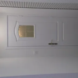 Białe drzwi z szybką i złotą klamką, po remoncie, na tle świeżo pomalowanej ściany i fragmentu ciemnej płytki. Widoczny włącznik światła i listwa sufitowa.