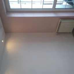 Jasne wnętrze po odświeżeniu: białe ściany, okno z widokiem na zewnątrz, grzejnik i ciemna podłoga z płytek. Akcent kolorystyczny w postaci różowego pasa pod oknem.