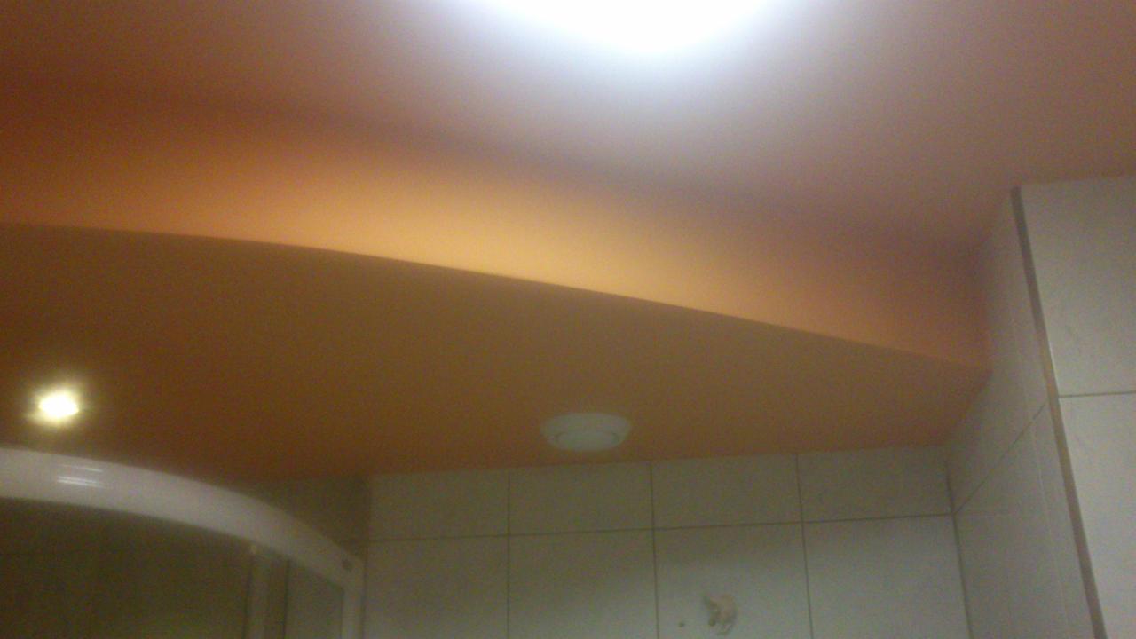 Pomarańczowy, gładki sufit w łazience z białymi płytkami i fragmentem kabiny prysznicowej. Widoczne oświetlenie sufitowe.