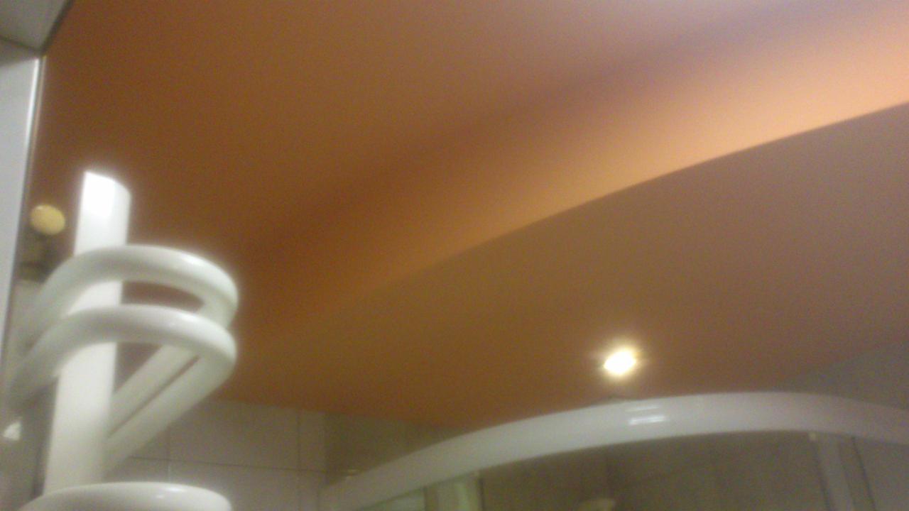 Pomarańczowy, gładki sufit napinany z oświetleniem punktowym w łazience, zrealizowany w Kleosin. Widoczny fragment kabiny prysznicowej i lampy.