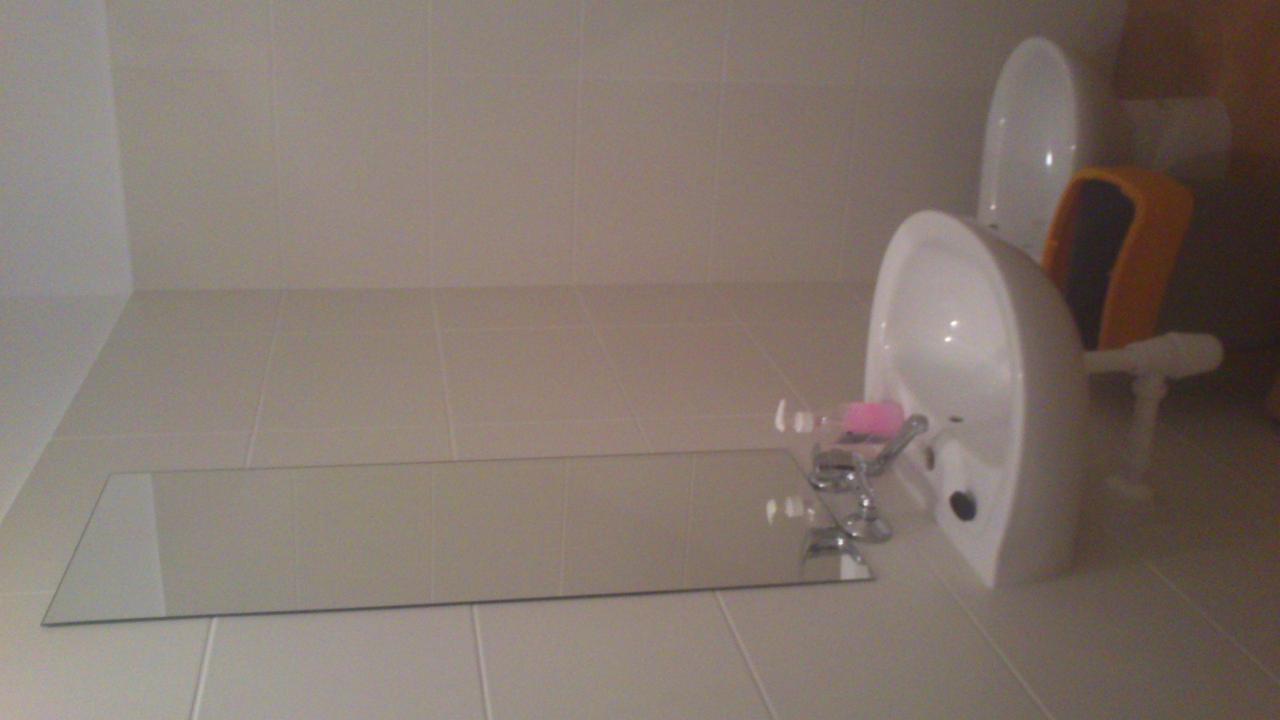 Ułożone płytki w łazience z umywalką i lustrem na podłodze. Widoczny fragment toalety i akcesoria łazienkowe. Jasne, neutralne kolory płytek.