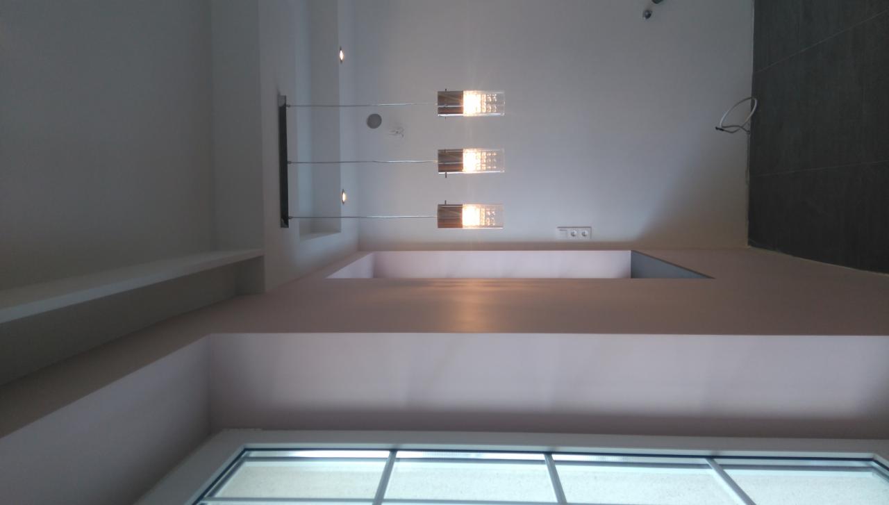 Nowoczesny sufit podwieszany z oświetleniem punktowym i lampą wiszącą. Widoczna także ściana wykończona płytkami i okno.