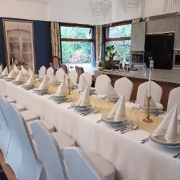 Uroczysty stół z białym obrusem, złotym bieżnikiem, nakrytymi miejscami z eleganckimi serwetkami, szklankami i sztućcami, oraz świecznikami; w tle nowoczesna kuchnia i widok na ogród.