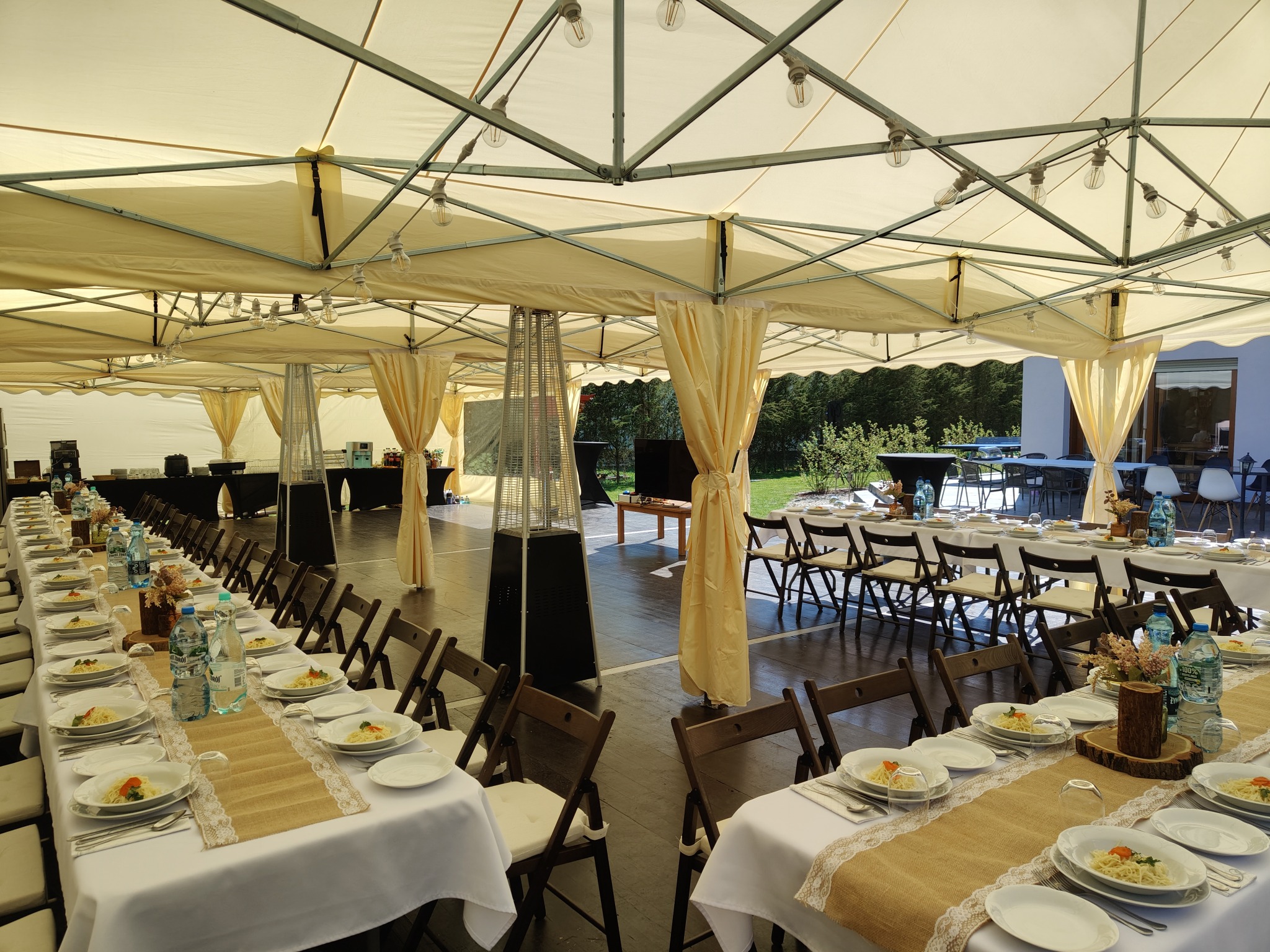 Długie stoły z białymi obrusami i dekoracjami, nakryte do obiadu pod kremowym namiotem z oświetleniem, przygotowane na przyjęcie.