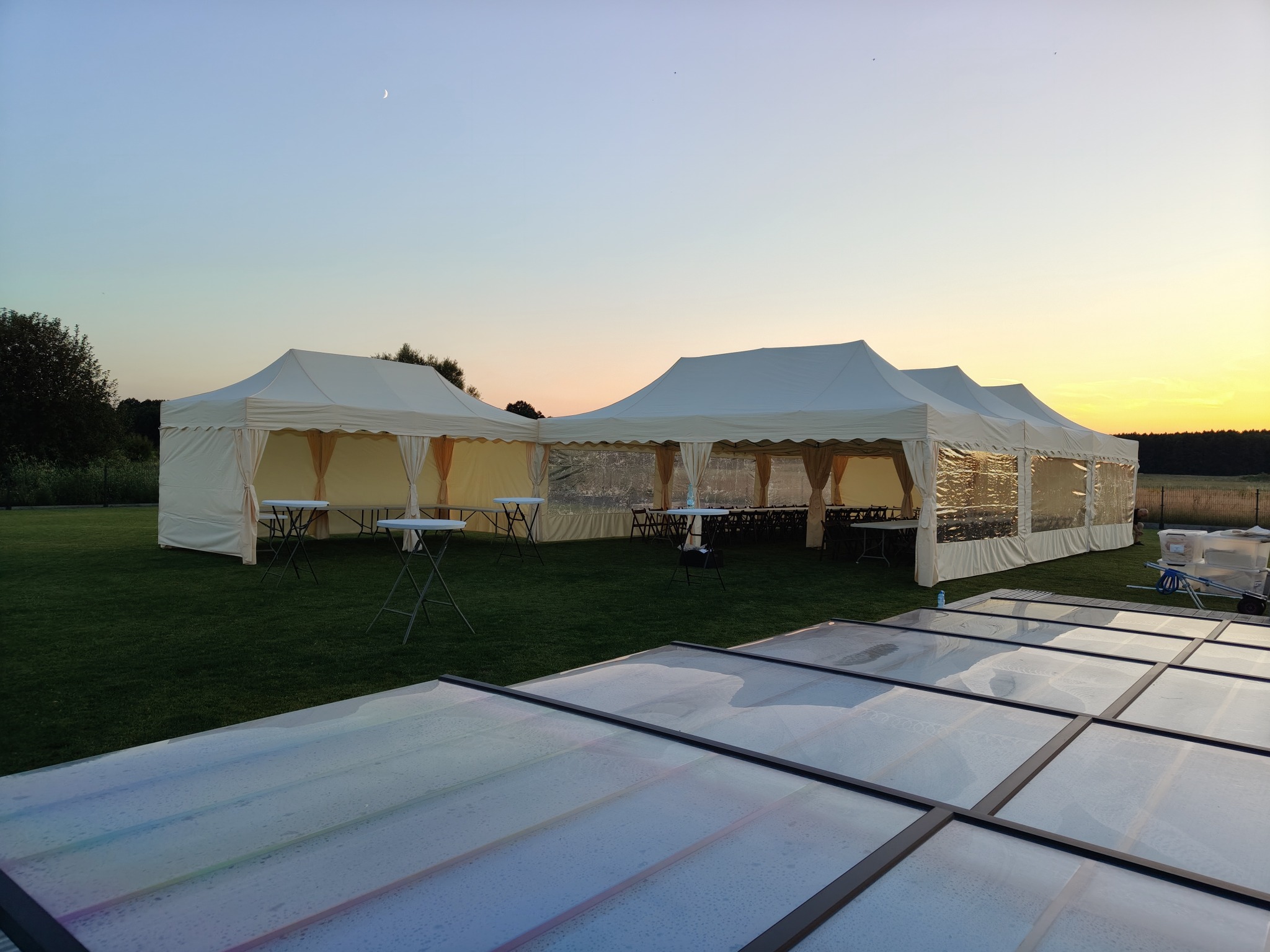 Rozstawione, połączone białe namioty cateringowe na zielonym trawniku, przygotowane do obsługi imprezy plenerowej o zmierzchu. Widoczne stoły i krzesła wewnątrz.