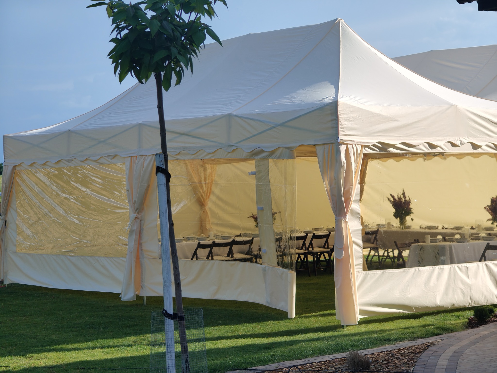 Biały namiot cateringowy na zielonym trawniku, widoczne stoły z krzesłami wewnątrz. Ustawiony na zewnątrz, gotowy na imprezę. Dekoracje kwiatowe na stołach.