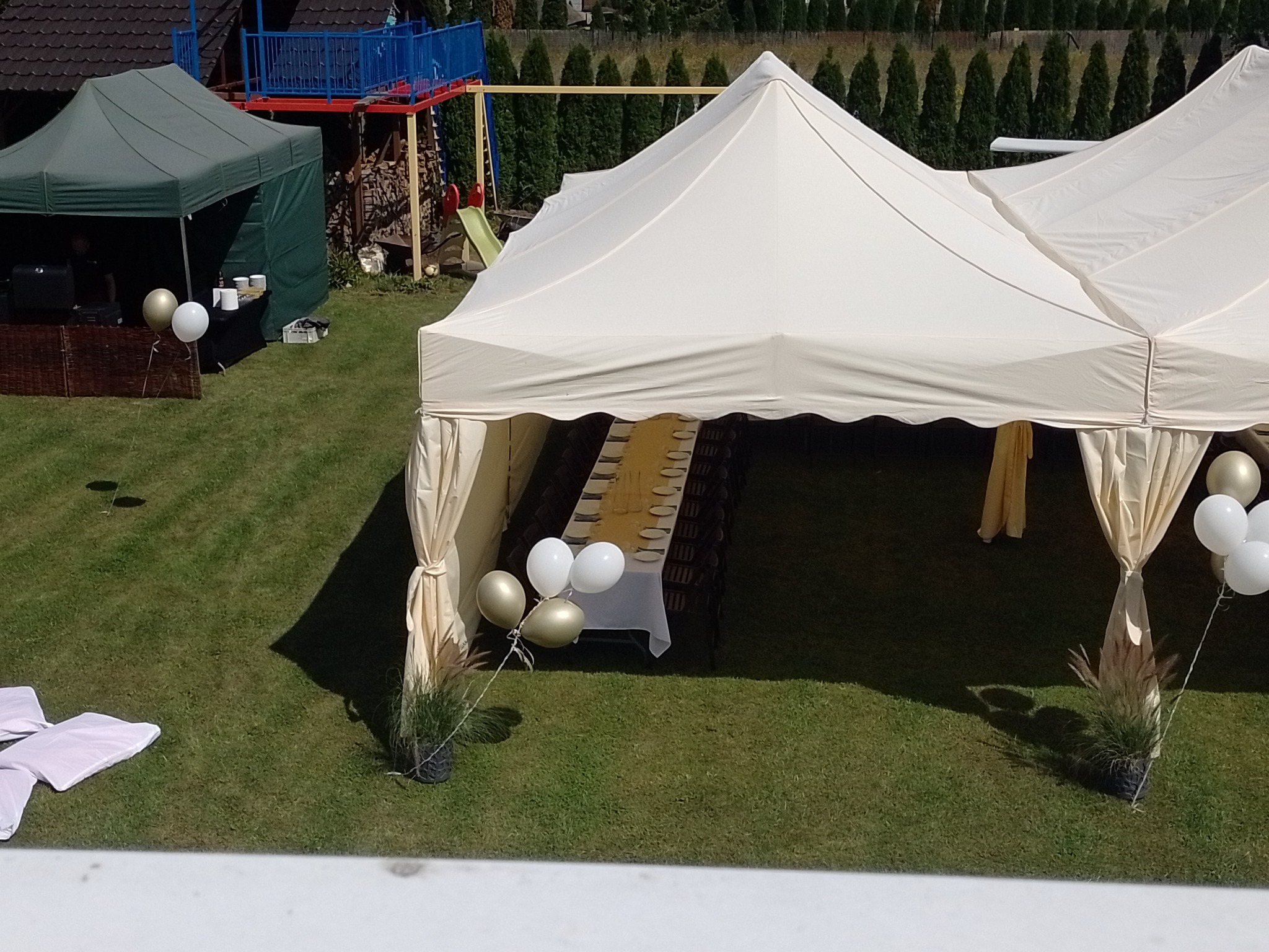Biały namiot cateringowy z zastawionym stołem na zewnątrz, dekorowany balonami i trawą w donicach, w tle zielony namiot i plac zabaw.