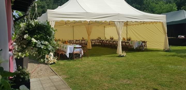 Uroczysty catering plenerowy: biały namiot, stoły z dekoracjami, krzesła i kwiaty na trawniku. Idealne na imprezę firmową lub świąteczną.