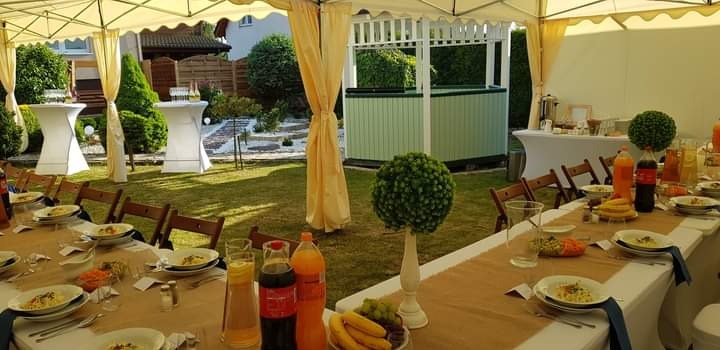 Uroczysty catering w plenerze: zastawiony stół pod namiotem, dekoracje roślinne, napoje i potrawy. W tle ogród z białymi podestami.