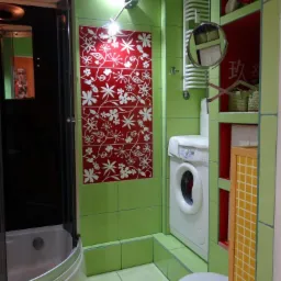 Jasnozielona łazienka z kabiną prysznicową, pralką i czerwonym panelem dekoracyjnym z białymi kwiatami. Oświetlenie sufitowe i biała armatura.