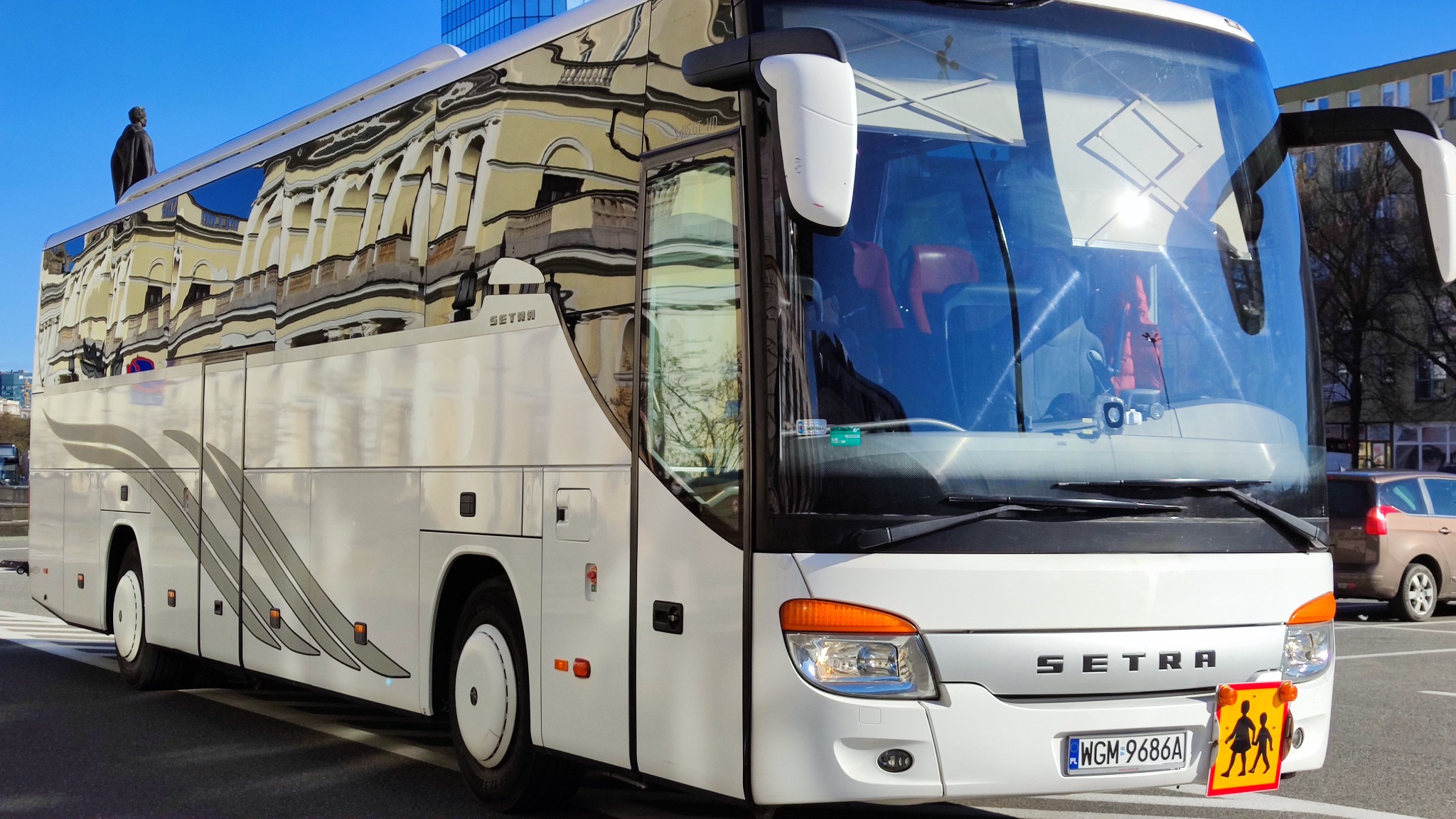 Biały autokar Setra z tabliczką informującą o przewozie dzieci, zaparkowany na parkingu z odbiciem budynku w szybach.
