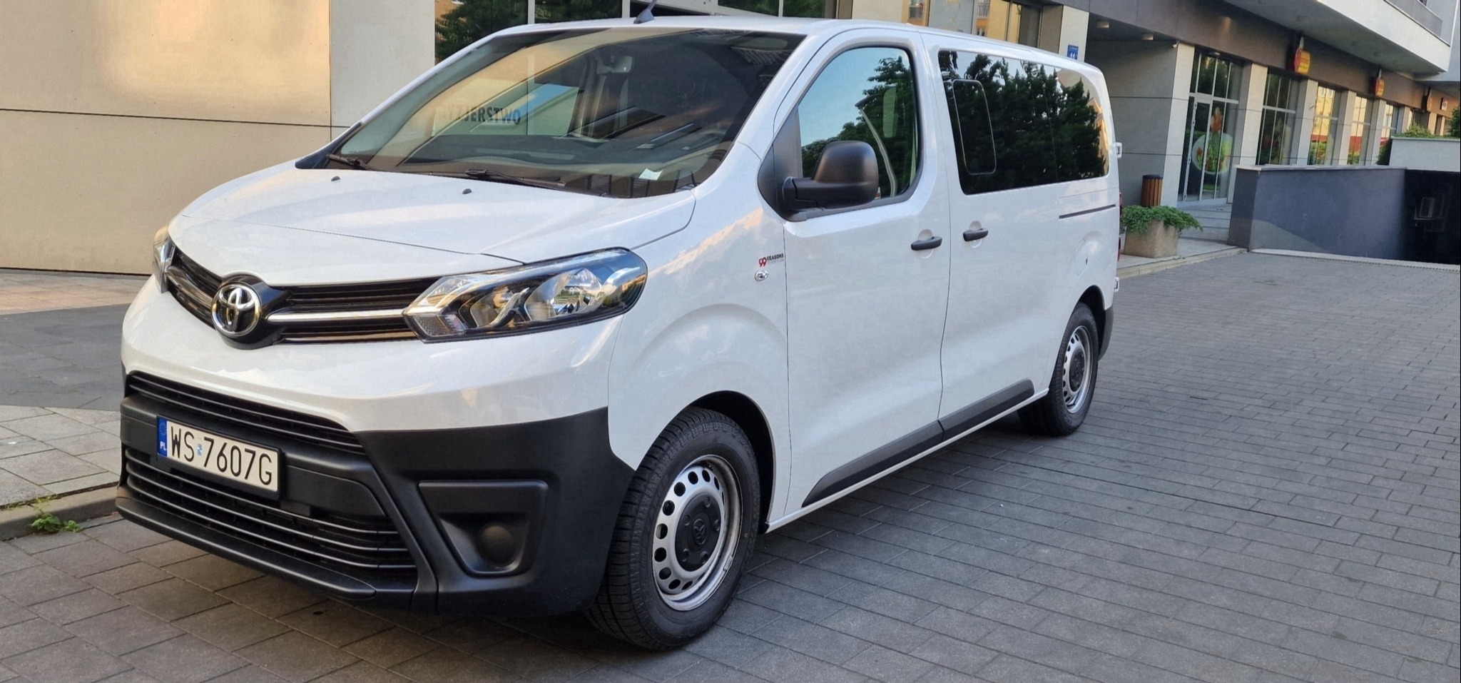 Biały bus Toyota Proace Verso z polską tablicą rejestracyjną WS 7607G, zaparkowany na chodniku przed budynkiem z restauracją.