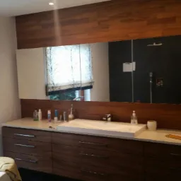 Nowoczesna łazienka z drewnianymi akcentami: długa szafka pod umywalką z szufladami, duże lustro z odbiciem kabiny prysznicowej, roleta w oknie z motywem drzewa.