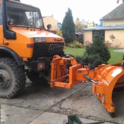 Pomarańczowy Unimog z pługiem śnieżnym na betonowym podjeździe, widok z boku, w tle budynki mieszkalne i zieleń. Tablica rejestracyjna: SZY 6HX5.