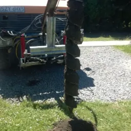 Wiertnica z wiertłem spiralnym, zagłębiona w ziemi na trawniku, z pomarańczowym ciągnikiem w tle. Widoczny gruz przy trawniku.