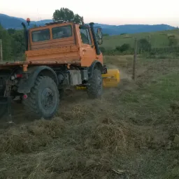 Pomarańczowy Unimog z glebogryzarką w trakcie pracy na polu. W tle widoczne wzgórza i drzewa. Maszyna w ruchu, unoszący się kurz.