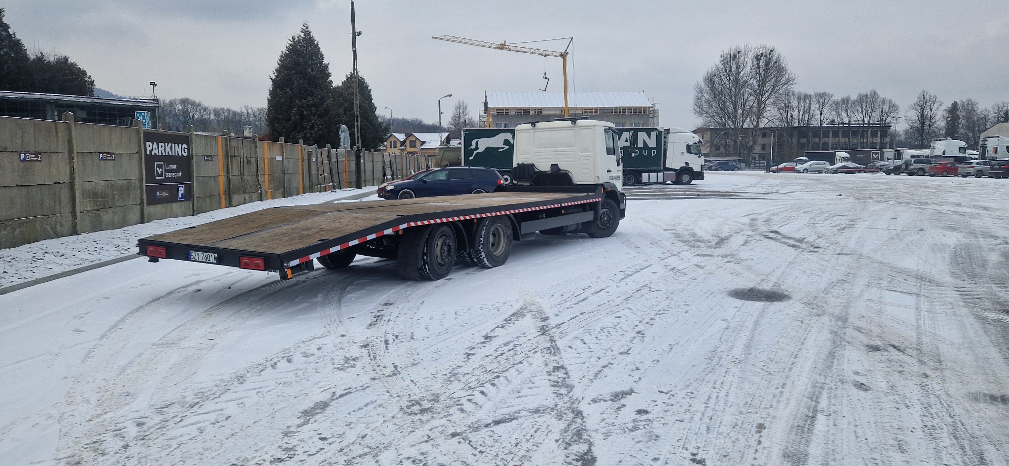 Laweta na parkingu w zimowej scenerii. W tle budynek w trakcie budowy z dźwigiem. Widoczne ślady opon na śniegu.