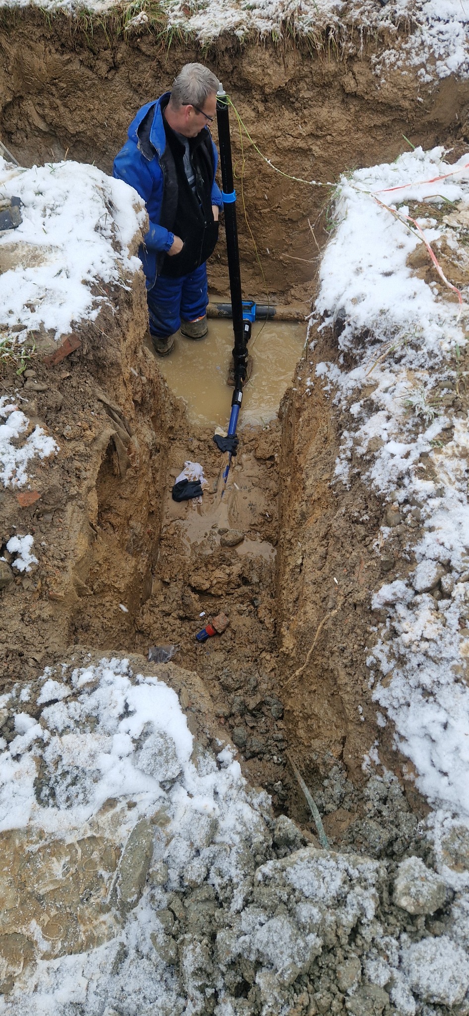 Pracownik w wykopie z wodą, instalujący rurę. Śnieg na ziemi, błoto i narzędzia w dole. Widok z góry na etap prac instalacyjnych.
