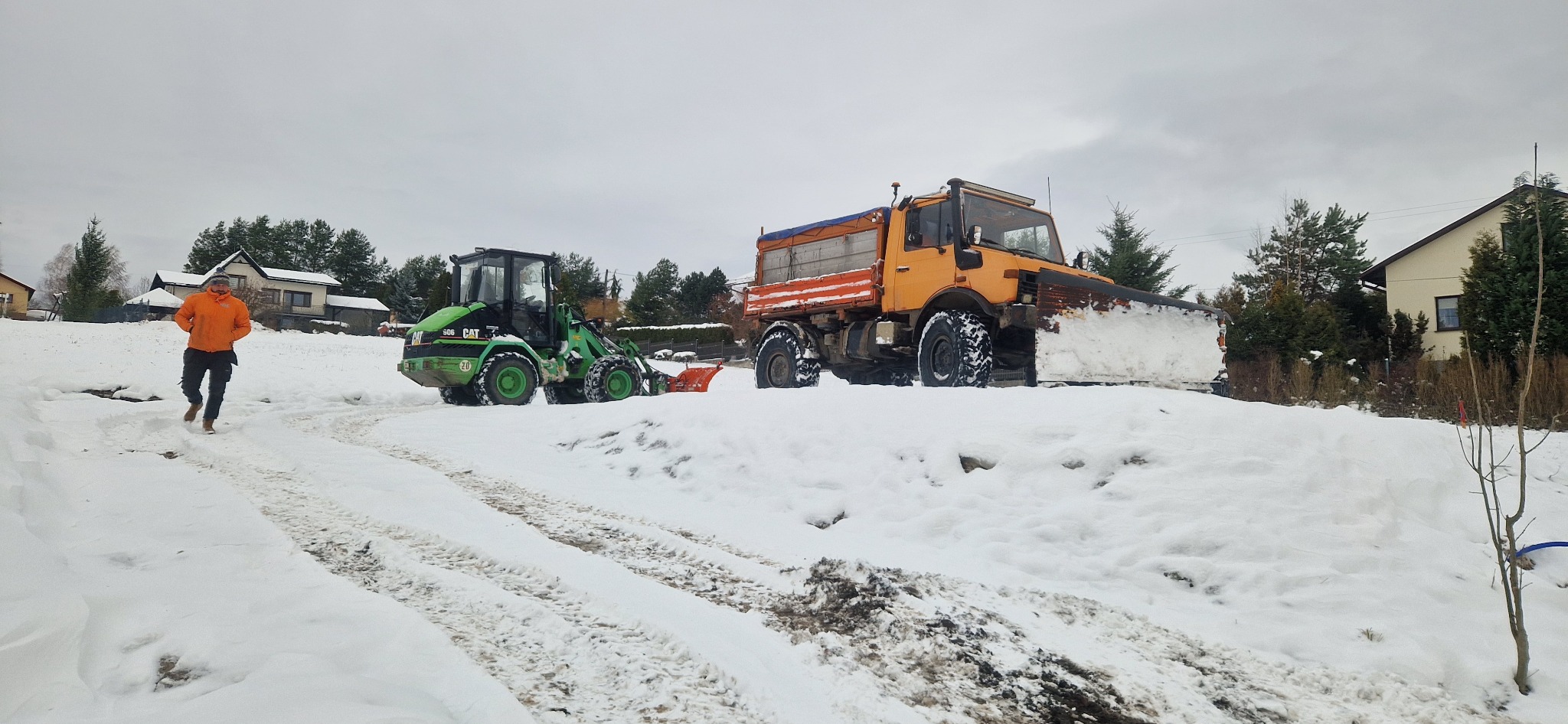 Odśnieżarka i mała ładowarka CAT na zaśnieżonej drodze, w tle domy i drzewa. Mężczyzna w pomarańczowej kurtce idzie w kierunku maszyn.