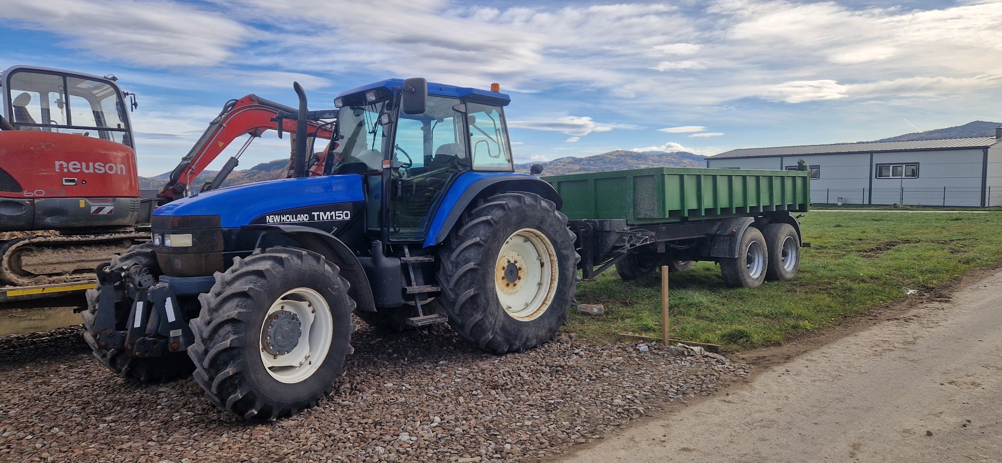 Niebieski traktor New Holland TM150 z zieloną przyczepą na żwirowym podłożu, w tle czerwona koparka i budynek. Krajobraz z górami i pochmurnym niebem.