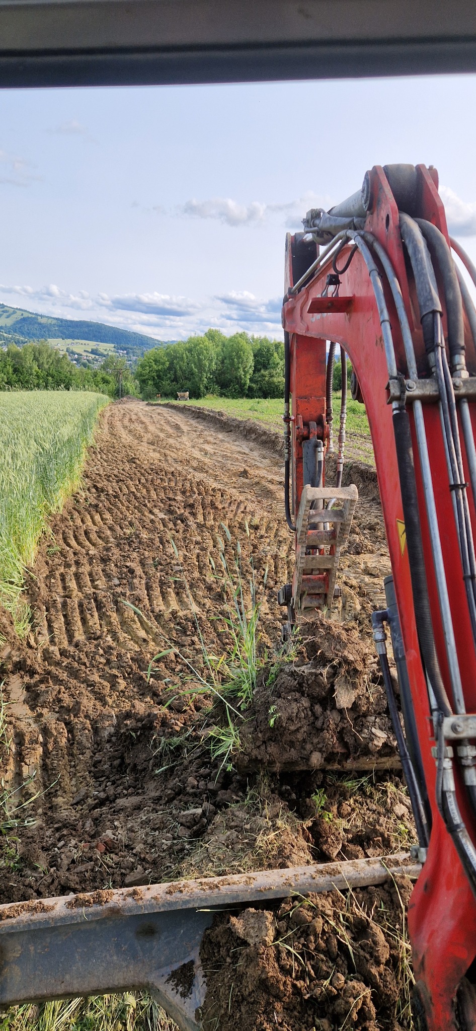 Czerwona koparka w trakcie prac ziemnych na polu, widok z kabiny operatora. W tle zielone wzgórza i błękitne niebo z chmurami.
