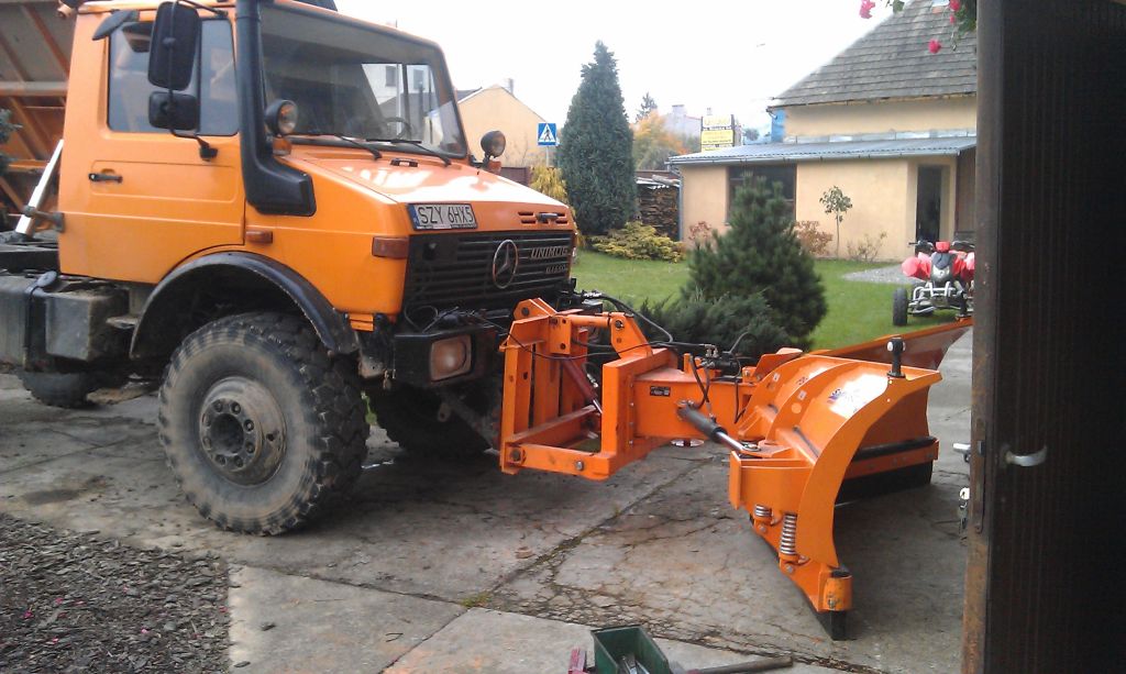 Pomarańczowy Unimog z pługiem śnieżnym na betonowym podjeździe, widok z boku, w tle budynki mieszkalne i zieleń. Tablica rejestracyjna: SZY 6HX5.
