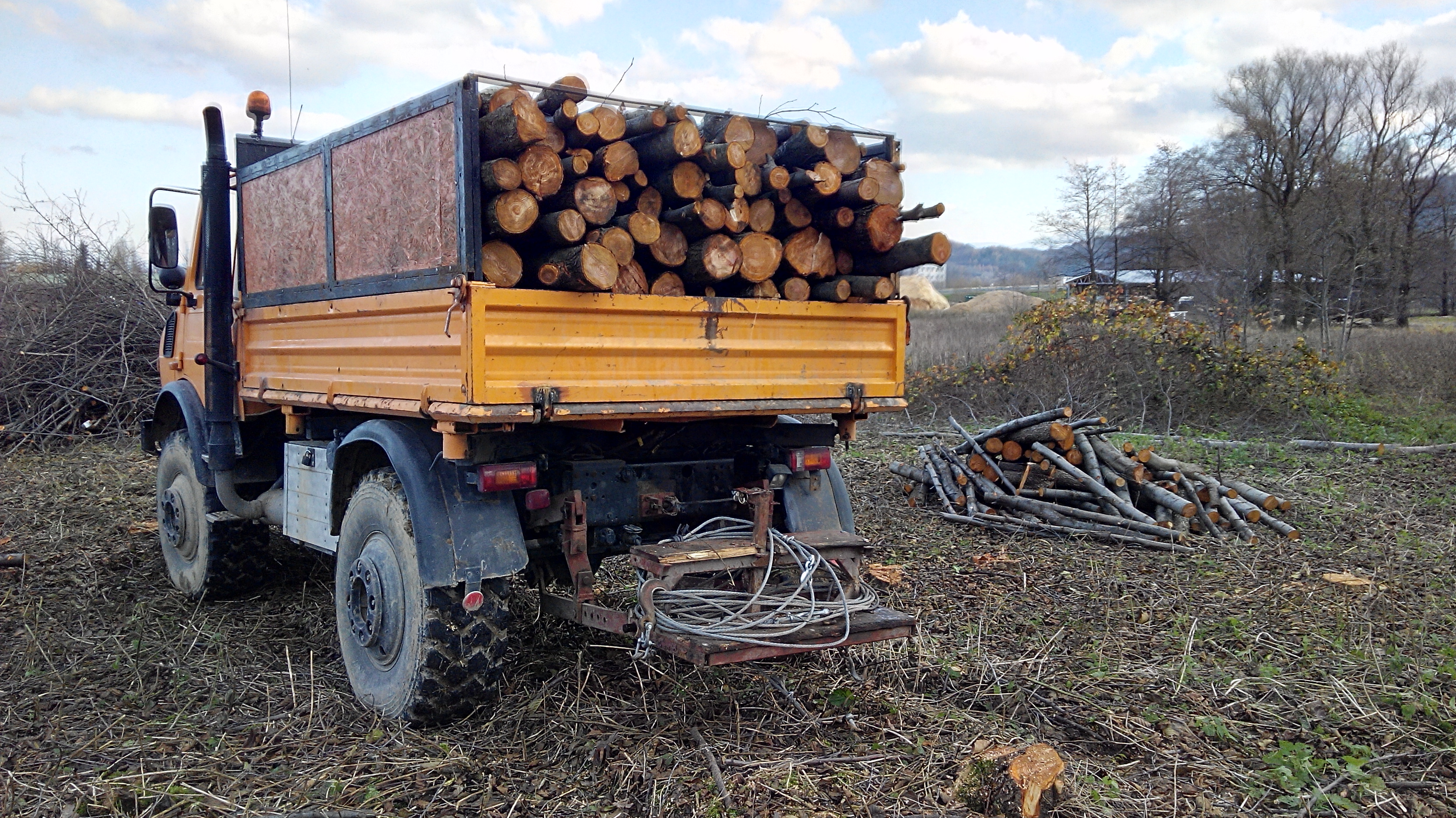 Pomarańczowy samochód ciężarowy z drewnem na pace, zaparkowany na polu. W tle widoczne drzewa i budynki.
