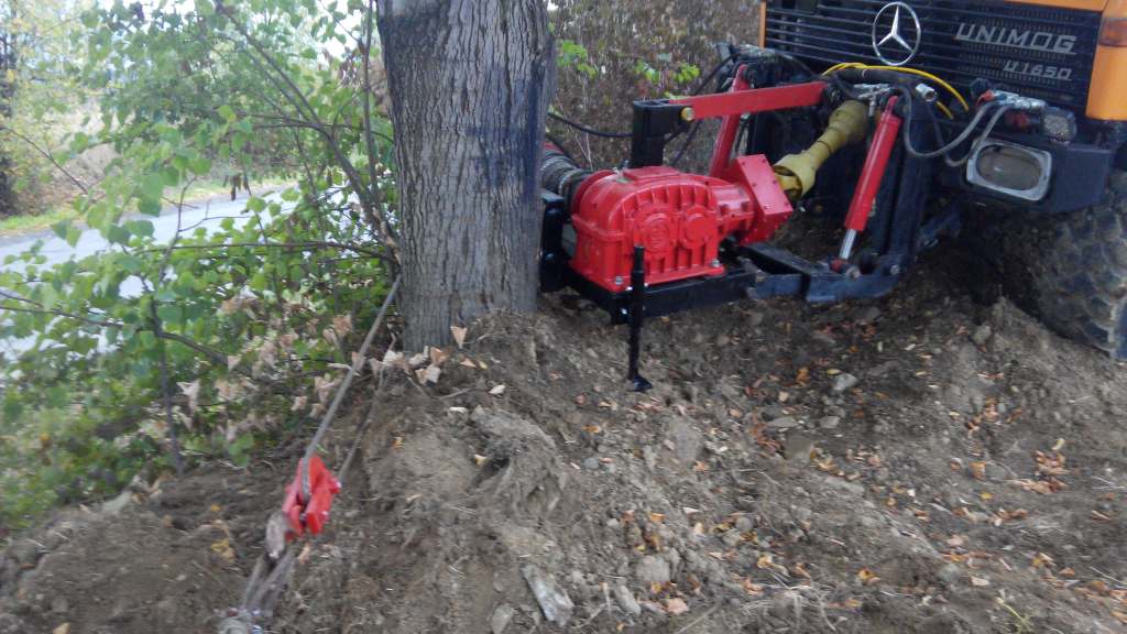 Unimog U1650 z wyciągarką linową, przymocowaną do drzewa, na nierównym terenie. Żółta karoseria, czerwona przekładnia, czarne elementy konstrukcyjne.
