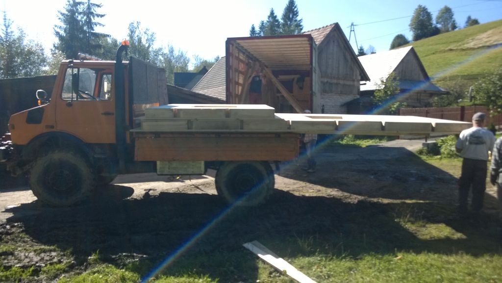 Pomarańczowy Unimog z platformą transportową na błotnistym terenie, w tle wiejskie zabudowania i wzgórze. Widoczni ludzie przy platformie.
