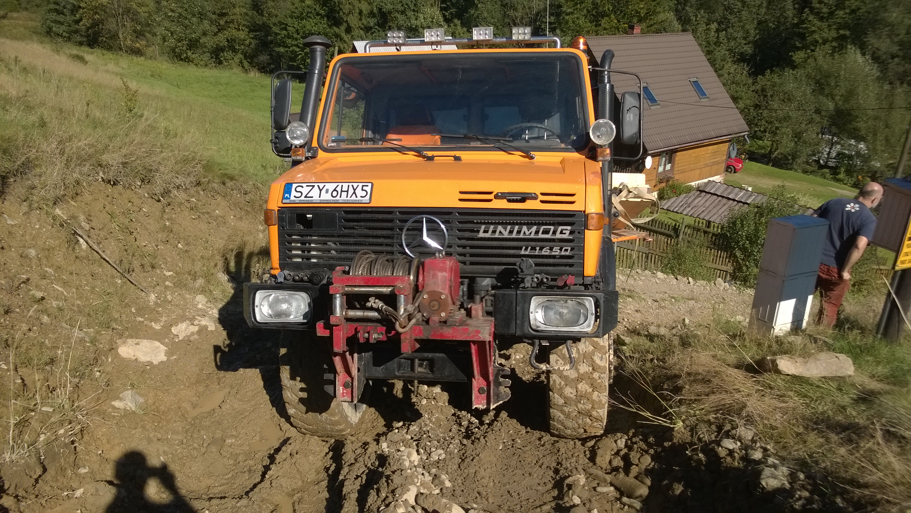Pomarańczowy Unimog U1650 z wyciągarką na błotnistym terenie, w tle zielone wzgórza i budynek. Widok z przodu, na pierwszym planie błoto i kamienie.