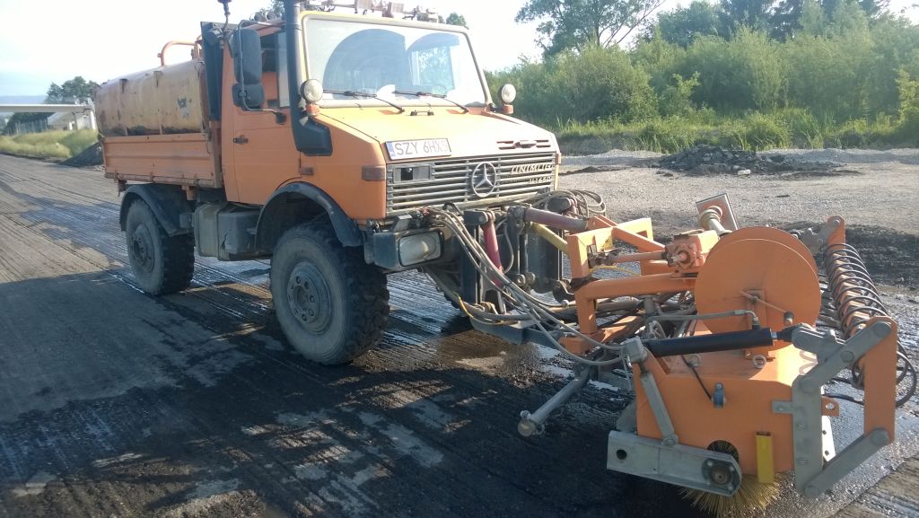 Pomarańczowy Unimog ze szczotką do czyszczenia dróg w trakcie pracy na asfalcie. Widoczny zbiornik na wodę i elementy hydrauliczne. Tło to zieleń i wiadukt.